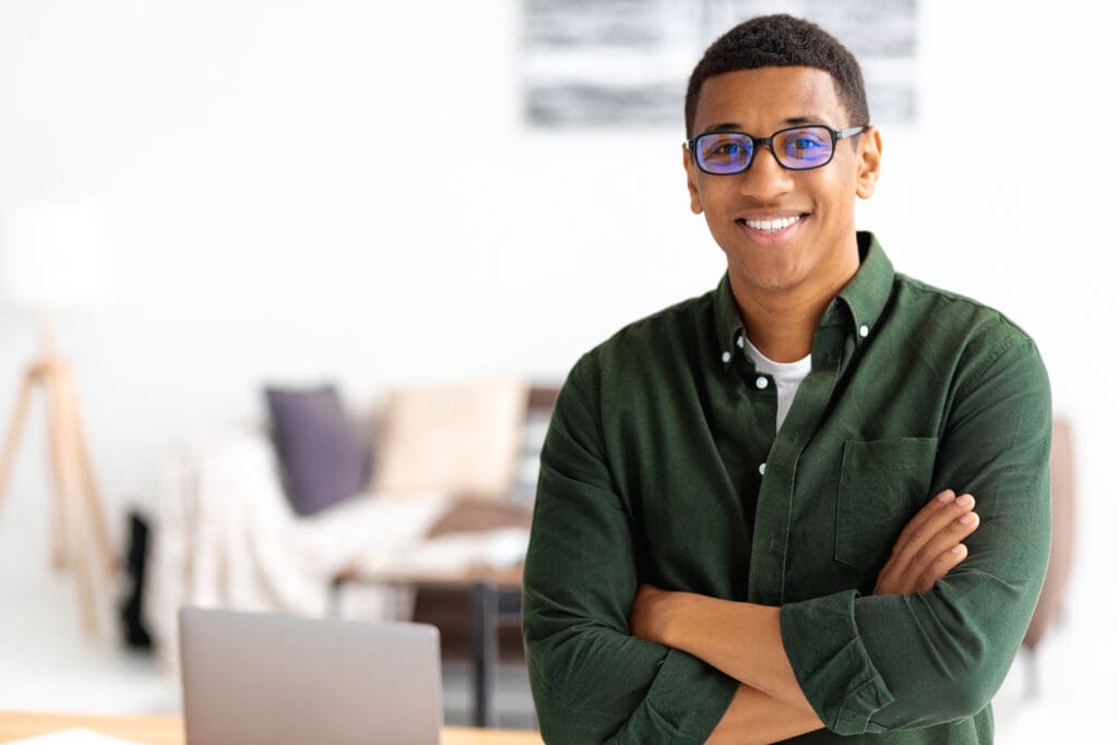 Smiling successful businessman with eyeglasses stands with crossed arms smiling and looking at the camera. Young male confident entrepreneur or small business owner in office